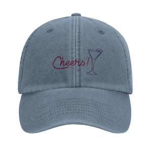 Könnte beinhalten: Blaue Baseballkappe mit dem gestickten Wort "Cheers!" in Schreibschrift, zusammen mit einer Cocktailglas-Illustration. Die Kappe hat einen gebogenen Schirm und einen Knopf oben.