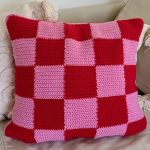 Op de afbeelding: Een vierkant gehaakt kussen met een rood en lichtroze dambordpatroon. Het kussen is handgemaakt en ziet er zacht en comfortabel uit, perfect voor de woondecoratie. Het ligt op een neutraal gekleurde bank.
