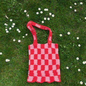 Könnte beinhalten: Handgefertigte Häkeltasche mit rot-rosa Schachbrettmuster. Die Tasche hat zwei rote Henkel und einen gewellten Rand. Die Tasche liegt auf grünem Gras mit kleinen weißen Blumen.