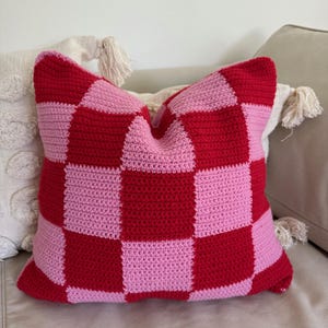Könnte beinhalten: Ein quadratisches Häkelkissen mit einem rot-rosa Schachbrettmuster. Das Kissen ist aus weichem Garn gefertigt und liegt auf einem neutralfarbenen Sofa. Ein dekoratives Wohnaccessoire.