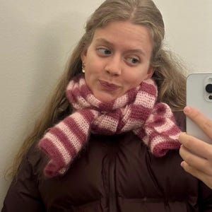 May include: A pink and white striped scarf, made of a soft, fuzzy material, is wrapped around the neck. The person in the image is wearing a dark maroon puffer jacket. The scarf's design features alternating stripes.