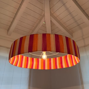 May include: A large, striped lampshade in shades of orange, pink, and beige hangs from the ceiling. The light fixture has a white metal frame and a single light bulb. The lampshade's design features vertical stripes.