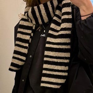 May include: A black and white striped scarf made of a soft, knitted fabric. The scarf is worn around the neck and is a classic winter accessory.
