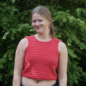 May include: A red and pink striped crochet top. The sleeveless top has a round neckline and a cropped fit. The crochet pattern features horizontal stripes of varying shades of red and pink. The top is worn by a person in front of a green leafy background.