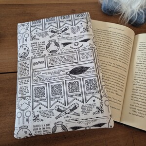 Può includere: Una copertina per libro in tessuto bianco con una stampa in bianco e nero di dettagli del gioco di Quidditch, tra cui il Boccino d'Oro, il Palla al Boccino, la Pluffa e le quattro case di Hogwarts: Grifondoro, Serpeverde, Tassorosso e Corvonero. Il testo include le regole del gioco e i nomi dei giocatori. La copertina è progettata per un libro di Harry Potter.