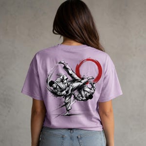 Peut inclure: Un t-shirt à manches courtes violet clair avec une impression graphique en noir et blanc au dos. Le motif représente deux personnages en uniforme d'arts martiaux effectuant un coup de pied dynamique, souligné par un cercle rouge.