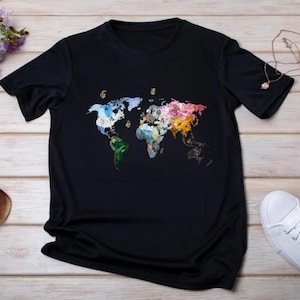 Peut inclure: T-shirt noir orné d'une carte du monde colorée. La carte est composée de diverses couleurs et motifs, chaque continent étant représenté par un style artistique unique. Le t-shirt est fait d'une matière douce.