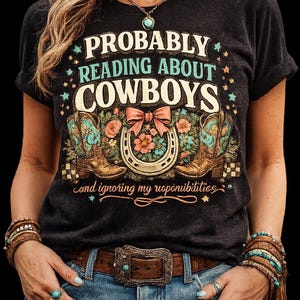 Peut inclure: Un t-shirt noir avec le texte PROBABLY READING ABOUT COWBOYS et and ignoring my responsibilities. Le motif comprend deux bottes de cow-boy marron, un fer à cheval, un nœud rose et des fleurs dans des tons de rose et de turquoise.