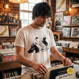 Peut inclure: Un t-shirt blanc à manches courtes avec une illustration graphique noire représentant deux personnages effectuant une projection d'arts martiaux. Le motif montre un personnage debout tandis que l'autre est en l'air, dans un mouvement circulaire.