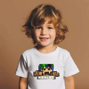 May include: A white t-shirt with a pixelated graphic of a character with green and blue hair, brown arms, and the word "ROBLOX" underneath. The shirt is worn by a young child with light brown hair.