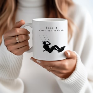 Könnte beinhalten: Ein weißer Keramikbecher mit einer schwarzen Silhouettengrafik einer Person beim Kitesurfen. Über der Grafik steht der Text: "home is where the wind blows." Der Becher verfügt über einen geschwungenen Henkel auf der linken Seite und eine glatte Oberfläche.