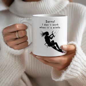 Könnte beinhalten: Ein weißer Keramikbecher mit einer schwarzen Silhouette-Grafik einer Person beim Kitesurfen. Der aufgedruckte Text auf dem Becher lautet: Sorry! I don't work when it's windy. Der Becher verfügt über einen Standardhenkel und eine glatte, glänzende Oberfläche.