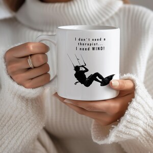 Könnte beinhalten: Ein weißer Keramikbecher mit einer schwarzen Silhouette-Grafik einer Person beim Kitesurfen, wobei die Leinen nach oben verlaufen. Der in Schwarz gedruckte Text lautet, I don't need a therapist... I need WIND! Der Becher hat einen Henkel.