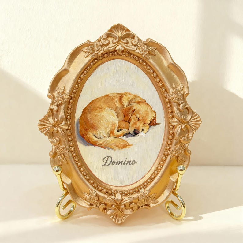 May include: Ornate gold-colored oval picture frame with a sleeping golden retriever painting. The frame has decorative floral and scroll details. The dog's name, "Domino," is written below the image. The frame is supported by a gold-colored stand.