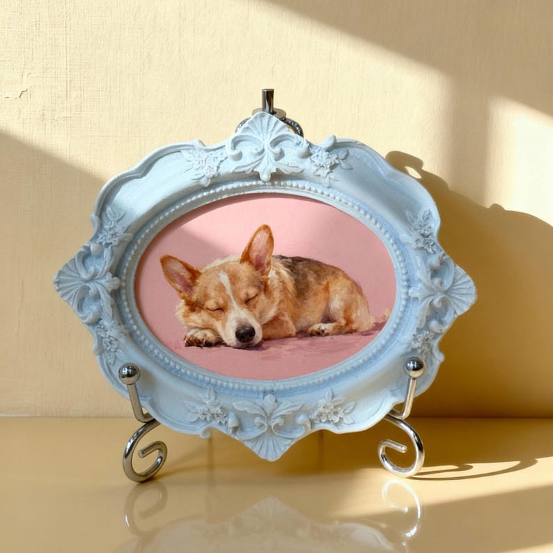 May include: A light blue, ornate picture frame with a silver stand. The frame holds an oval-shaped image of a sleeping corgi dog on a pink background. The dog has brown and white fur.