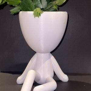 Può includere: Vaso da fiori in ceramica bianca a forma di persona seduta con le gambe incrociate. Il vaso ha una parte superiore larga e rotonda e una finitura liscia e opaca. Una pianta verde sta crescendo all'interno del vaso.