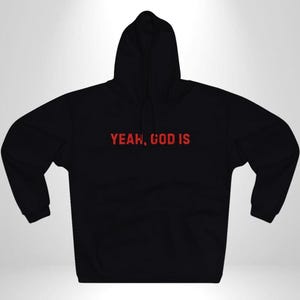 Op de afbeelding: Zwarte hoodie met een capuchon en een voorzak. De rode tekst "YEAH, GOD IS" is over de borst gedrukt. De hoodie is gemaakt van een zacht materiaal.