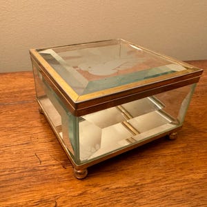 May include: A square glass trinket box with a gold metal frame and feet. The top of the box has a frosted design.