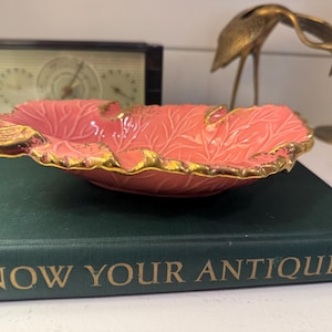 May include: A coral-colored, leaf-shaped ceramic dish with gold trim. The dish is resting on a green book titled "KNOW YOUR ANTIQUES". A vintage clock and decorative gold bird figurines are in the background.