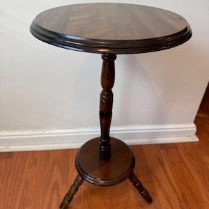 May include: A small, round, dark brown wooden side table. The table has a circular top with a slightly raised edge, supported by a turned pedestal and three legs. The table is approximately 24 inches tall.