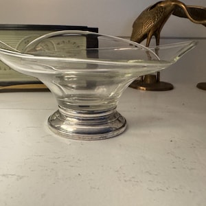 May include: A clear glass serving dish with a silver-colored base. The dish has a wide, shallow bowl and flared sides. Decorative brass bird figurines are in the background. The dish is likely vintage and suitable for serving snacks or small items.