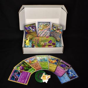 May include: A white cardboard box filled with Pokémon trading cards, a Pikachu figurine, and a camouflage-patterned tin. Several cards are displayed, including a green Bulbasaur card and a purple Gengar card. The box sits on a black surface.