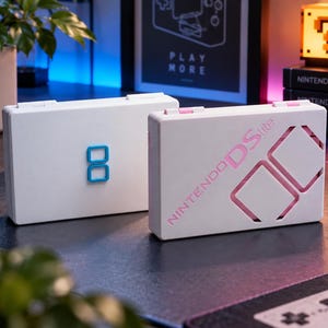 以下が含まれることがあります： 2台の白いNintendo DS Lite携帯ゲーム機。左側の本体には青いDSロゴが配置されています。右側の本体にはピンク色の「NINTENDO DS lite」という文字と、内部のピンク色の表面が見える2つの四角い切り抜きが施されています。