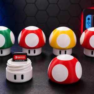 May include: A collection of colorful mushroom-shaped game cartridge holders. The holders are red, green, and yellow with white spots. One holder contains a Nintendo Switch game cartridge. The items are on a dark surface.