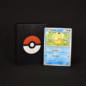Pu&ograve; includere: Un portamazzo rettangolare nero con un emblema Pok&eacute; Ball rosso e bianco, accanto a una carta collezionabile Pok&eacute;mon di Psyduck con 70 HP, che mostra gli attacchi Overthink e Water Gun, su uno sfondo nero uniforme.