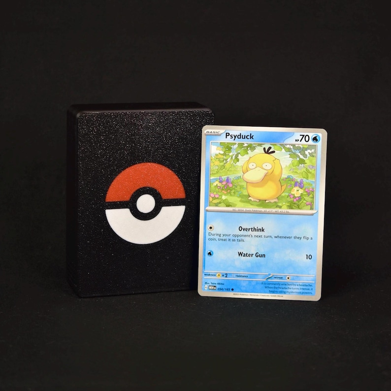 Pu&ograve; includere: Un portamazzo rettangolare nero con un emblema Pok&eacute; Ball rosso e bianco, accanto a una carta collezionabile Pok&eacute;mon di Psyduck con 70 HP, che mostra gli attacchi Overthink e Water Gun, su uno sfondo nero uniforme.