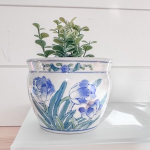 May include: A white ceramic planter with blue floral designs and green foliage. The planter is sitting on top of two books with the titles "Entertaining by" and "Waterside Cottages". The planter is a decorative item for home decor.