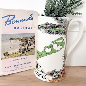 May include: A white ceramic mug with a Bermuda map design and a handle, holding a small green plant. Beside it is a vintage travel guide titled "Bermuda Holiday" with a pink cover. The mug features a map of Bermuda and palm tree illustrations.