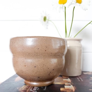 May include: A speckled tan ceramic bowl with a rounded body and a small footed base. The bowl has a smooth, glossy finish and sits atop a stack of two books, one titled Laying the Elegant Table and the other Litchfield St, with white daisies.