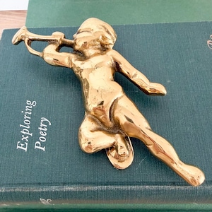 May include: A brass figurine of a cherub playing a trumpet. The cherub is kneeling with one leg extended and the other bent. The figurine is resting on a green book with the words "Exploring Poetry" on the cover.