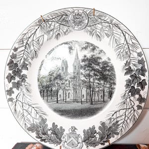 May include: A white ceramic decorative plate featuring a black and white transfer print of a church building surrounded by trees. The border displays pine branches, ivy leaves, a sun emblem with the text Bowdoin College, and a heraldic crest.