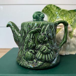 May include: A green ceramic teapot with a detailed relief of two rabbits in a forest setting. The teapot has a handle, spout, and a lid with a small rabbit on top. The teapot is sitting on a green book.