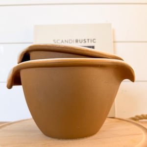 May include: A brown ceramic mixing bowl with a curved handle, sitting on a wooden cutting board. The bowl has a wide rim and a smooth, matte finish. A book with the text "SCANDIRUSTIC" is in the background.