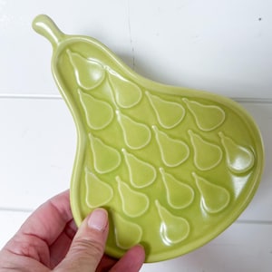 May include: A light green pear-shaped ceramic dish with a stem. The dish has a series of pear-shaped indentations on its surface. The dish is held against a white background.