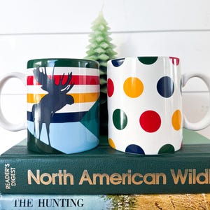 May include: Two white ceramic mugs. One mug features a moose silhouette against a colorful striped background. The other mug has a white base with large, colorful polka dots. Both mugs have white handles.