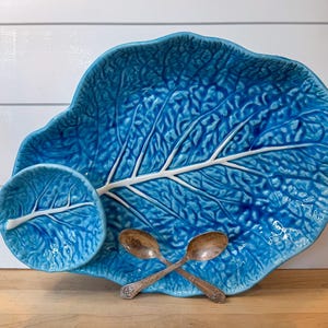 May include: A large, turquoise ceramic serving platter and a small matching dish, both shaped like leaves with white vein detailing. Two antique silver spoons are crossed in front of the dishes. The background is a white wooden wall.
