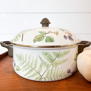 May include: Cream-colored enamelware pot with lid, featuring a botanical design of green ferns, leaves, and purple berries. The pot has dark handles and a gold rim. The lid is topped with a brown knob. A decorative kitchen item.