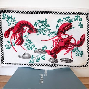 May include: White kitchen towel with a black and white checkered border, featuring two red lobsters, green sprigs, and gray seashells. The towel is folded over a blue book titled "Waterside Cottages".