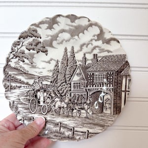 May include: A decorative plate with a scalloped edge featuring a sepia-toned pastoral scene. The image depicts a horse-drawn carriage, a building, trees, and a fence. The plate is held by a hand against a white background.