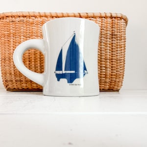 May include: A white ceramic mug featuring a blue graphic of a sailboat on the front. The design includes the text ©2001 Dan Morris printed in small black letters at the base of the sailboat illustration. The mug has a curved handle on the left side.
