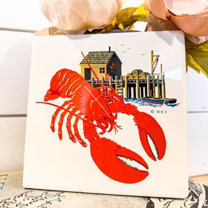 May include: Square ceramic tile featuring a vibrant red lobster illustration. The tile also depicts a coastal scene with a small building, pier, and boat. The tile is white with a glossy finish, perfect for kitchen or coastal decor.