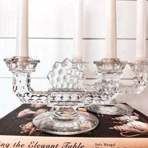 May include: A clear glass candelabra with four candle holders, each holding a white candle. The candelabra sits atop a stack of books, including one titled "ing the Elegant Table". The candelabra has a geometric design.