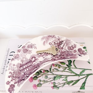 May include: A crescent-shaped ceramic dish featuring a purple and white transferware pattern of a landscape with buildings and flowers. A single brass-colored metal key rests in the center of the dish, which has a scalloped edge design.