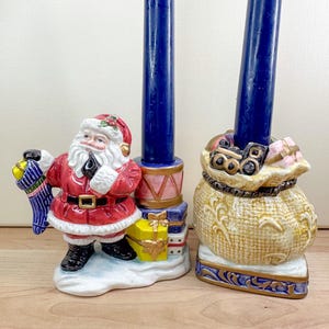 May include: Two Christmas-themed candle holders. One features Santa Claus holding a stocking, the other a sack of toys. Both have tall, dark blue candles. The holders are ceramic with colorful details, including a gift box.