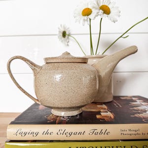May include: A speckled beige ceramic teapot with a lid, a curved handle, and a long spout. The teapot rests on two stacked books titled Laying the Elegant Table and Litchfield S. Behind the teapot, three white daisies with yellow centers are in a vase.
