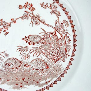 May include: A white ceramic plate featuring a detailed red transferware pattern. The design depicts two small birds foraging near flowering branches, bamboo, and large blossoms. A decorative geometric border frames the edge of the plate.